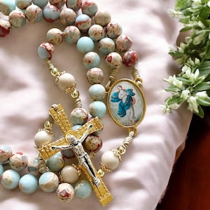 May include: A blue and brown stone rosary with a gold cross and a silver image of the Virgin Mary. The rosary is strung on a beige cord.