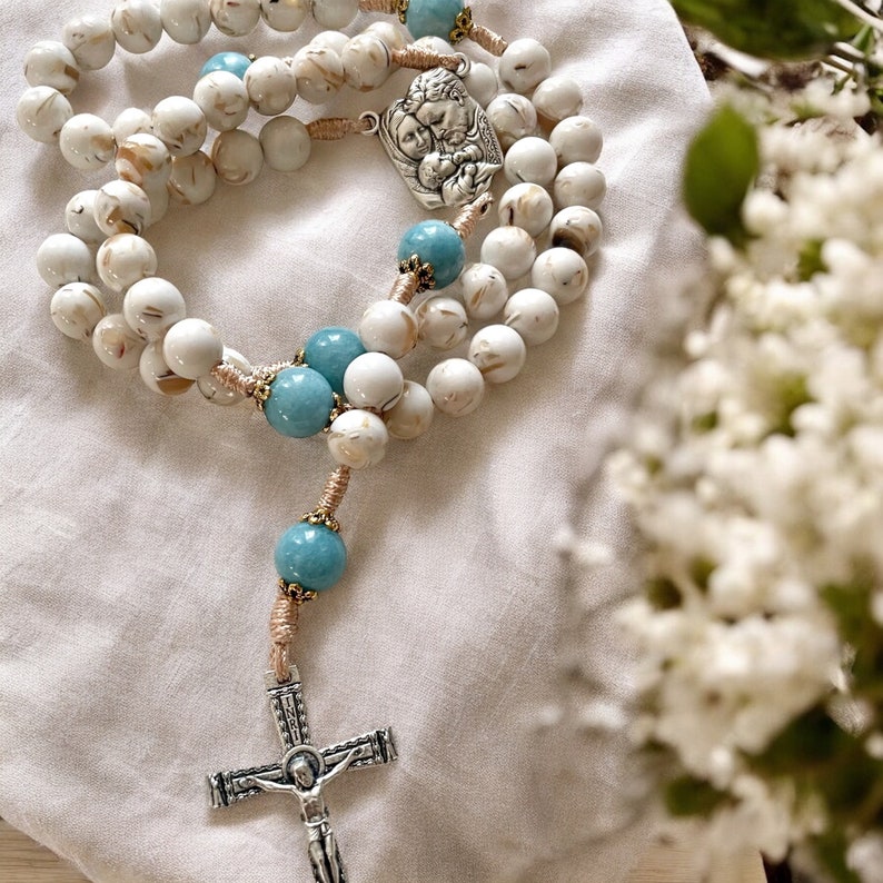 Puede incluir: Rosario de cuentas blancas y azules con un colgante de plata que representa a la Sagrada Familia. Las cuentas son de piedra blanca con detalles azules.