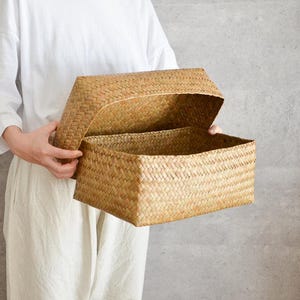 以下が含まれることがあります： 長方形の織りバスケットで、蓋は開いています。バスケットは天然色の織り素材で作られています。蓋は開いており、内部が見えます。バスケットはニュートラルな灰色の背景に対して保持されています。