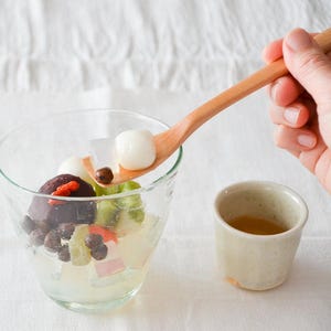 Könnte beinhalten: Eine Glasschale gefüllt mit japanischen Dessert-Zutaten, darunter Mochi-Bällchen, rote Bohnenpaste und Obst. Ein Holzlöffel hält eine Mochi-Kugel über der Schale. Daneben steht eine kleine Keramiktasse Tee.