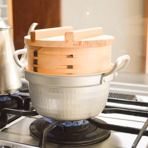 以下が含まれることがあります： 木製のハンドルが付いたステンレス製のケトルが、ガスコンロの横にある伝統的な木製の蒸し器の隣にあります。蒸し器は明るい色の木材と金属でできており、鍋の上に置かれています。コンロは点火されており、青い炎が見えます。