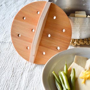 Peut inclure: Un couvercle de cuiseur vapeur en bois percé de trous est posé à côté d'une casserole en métal et d'une assiette blanche contenant du tofu, des haricots verts et une garniture jaune. Le couvercle est brun clair.