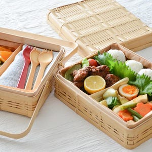 May include: Woven bamboo bento boxes, one open and filled with fruit and utensils, the other with a variety of foods. The open box contains strawberries, kiwi, orange slices, and wooden utensils. The other box contains rice balls, fried food, and vegetables.