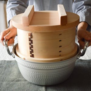 May include: A light-colored wooden steamer with a lid and a metal pot. The steamer has a rectangular handle on top and is held by a person. The pot has a textured surface and silver handles. The items are on a table.