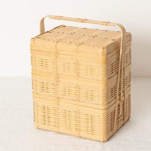 May include: A light brown, woven bamboo picnic basket with a rectangular shape and a handle. The basket is made of woven bamboo strips and has a lid. The handle is arched and made of the same material.