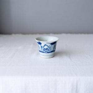 Peut inclure: Une petite tasse en céramique blanche avec des motifs floraux bleus. La tasse a un bord évasé et un corps légèrement conique. Le motif bleu présente des feuilles et des lignes stylisées. La tasse est posée sur une surface blanche.