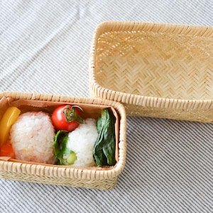 Può includere: Due scatole per il pranzo in bambù intrecciato, una aperta e una con coperchio. La scatola aperta contiene polpette di riso, un pomodoro rosso, fette di peperone giallo e verdure a foglia verde. Le scatole sono su un tessuto a righe.