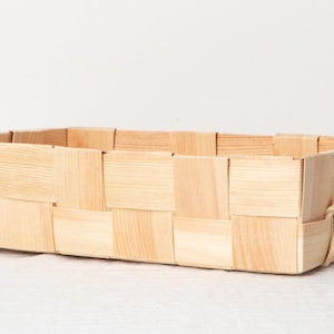May include: A rectangular woven basket made of light-colored wood. The basket features a woven pattern and has handles on each end. The basket is empty and sits on a white surface.