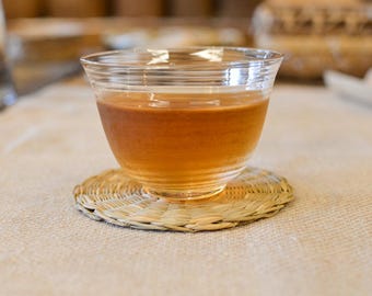 May include: A glass teacup containing amber liquid, placed on a woven straw coaster. The cup has a rounded shape and sits on a circular coaster. The background is blurred, suggesting a natural setting.