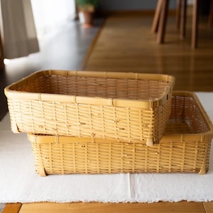 May include: Two rectangular woven bamboo baskets stacked on top of each other. The baskets are a light tan color and have a simple, open weave design. They are sitting on a white cloth.