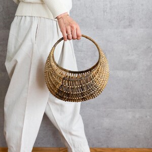 以下が含まれることがあります： 半月型の織りバスケットと円形の持ち手。バスケットは天然の葦で作られており、明るい茶色から濃い茶色へのグラデーションがあります。クリーム色の服を着た人がバスケットを持っています。