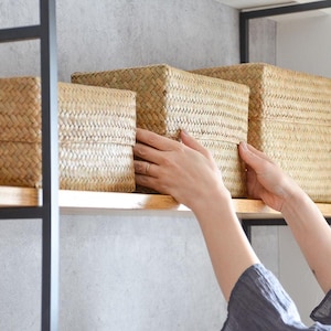 Può includere: Tre scatole portaoggetti intrecciate di diverse dimensioni sono su una mensola di legno. Le scatole sono realizzate in un materiale marrone chiaro e strutturato e hanno coperchi. Le mani di una persona raggiungono la scatola centrale.