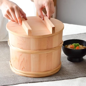 May include: A light-coloured wooden rice tub with a lid, cylindrical in shape, with a woven band around the middle. Two bowls of soup are next to the tub. A person is handling the tub.