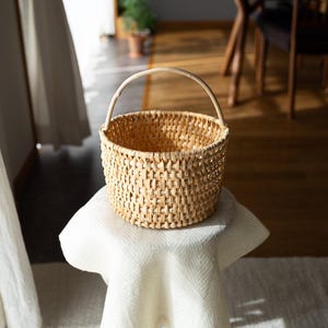 May include: A round, woven basket with a handle, made of light brown material. The basket is sitting on a white surface, with a window and wooden floor in the background. The basket is empty.
