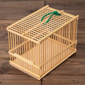 Peut inclure: Une petite cage à oiseaux rectangulaire en bois clair. La cage a des barreaux verticaux et un toit à lattes, avec une poignée en ficelle verte. La cage est posée sur une surface en bois.