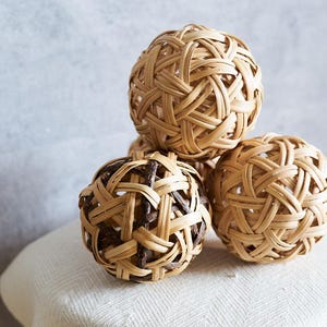 May include: Four woven rattan balls in a natural tan colour are stacked on a white surface. The balls have an intricate woven design, with one ball having darker brown accents. The background is a light grey.