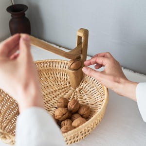 以下が含まれることがあります： 木製のくるみ割り器が使用されており、くるみがたくさん入った編みかごの上に配置されています。 くるみ割り器は薄茶色で、かごは自然なベージュ色です。 ニュートラルな背景の室内シーンです。