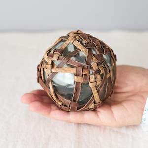 May include: A clear glass sphere enclosed in a woven basket made of brown and tan leather or rattan. The decorative ball is held in a hand, highlighting the intricate weave and the transparency of the glass within.