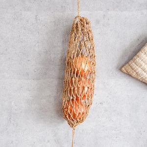 May include: A woven jute net bag filled with several brown onions hangs against a gray wall. A small, square, beige quilted item hangs from a metal hook to the right. The net bag is suspended by a jute cord.