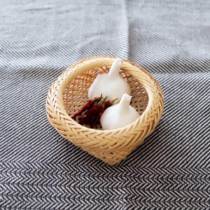 以下が含まれることがあります： 2つの白いニンニクの球根と、いくつかの乾燥した赤い唐辛子が入った、織られたバスケット。 バスケットは薄茶色の織り素材でできており、グレーと白のストライプの生地の上に置かれています。