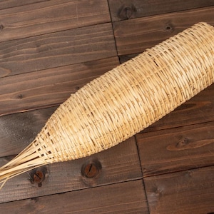 Può includere: Un cesto intrecciato di forma lunga e affusolata, realizzato con fibre naturali marrone chiaro. Il cesto è appoggiato su una superficie di legno marrone scuro.