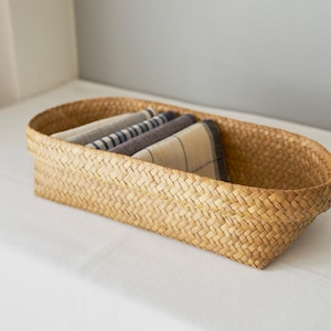 May include: An oval-shaped woven basket made of light brown natural fibers. The basket contains folded textiles in various neutral colors and patterns. The basket is sitting on a white surface.