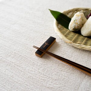 以下が含まれることがあります： 緑の葉が付いた編みかごに入った2つのご飯のおにぎりが特徴の日本の料理の盛り合わせ。木製の箸置きと箸が前景にあります。テーブルは白いテクスチャード加工の布で覆われています。