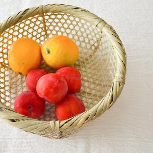 以下が含まれることがあります： 新鮮な果物が入った丸い竹製のバスケット。バスケットにはオレンジ2個と赤いプラム5個が入っています。バスケットは薄茶色で、白い表面に置かれています。