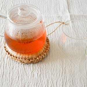 以下が含まれることがあります： 琥珀色の紅茶が入った透明なガラス製のティーポット。蓋と持ち手が付いており、編み込まれた藁の丸いコースターの上に置かれています。ティーポットの右側には、白い布の上に空の透明なガラス製カップが2つ並んでいます。