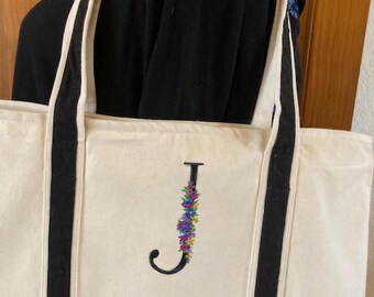 May include: A cream-colored canvas tote bag featuring a black base and two black shoulder straps. The front of the bag displays a large, embroidered letter J, which is decorated with a colorful floral pattern in shades of blue, purple, and yellow.