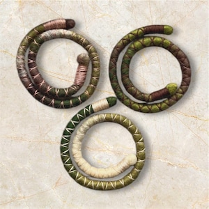 Op de afbeelding: Drie handgemaakte armbanden gemaakt van geweven garen in tinten groen, bruin en crème. De armbanden zijn opgerold in een cirkelvorm.
