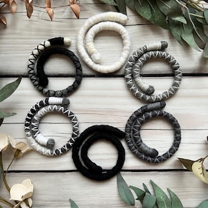 May include: Six coiled wool rope bracelets in black, white and grey. The bracelets are arranged in a circle on a white wooden surface.