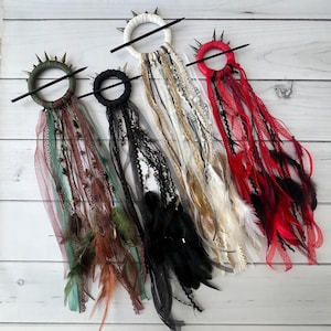 以下が含まれることがあります： 羽根とリボンが付いたヘアクリップが4つ。各クリップには黒い棒とスパイクのある円形のベースがあります。リボンは赤、白、黒、緑など、さまざまな色があります。