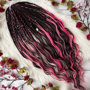 May include: A set of dark brown and black dreadlocks with vibrant pink highlights. The dreadlocks are styled with a mix of textures and adorned with small beads and charms. The hair is arranged on a white, fluffy surface, decorated with floral accents.
