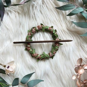 May include: A brown wooden hair stick with a green and brown floral wreath design. The wreath is made of faux leaves and twine.