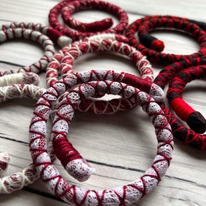 以下が含まれることがあります： レースのディテールが付いた赤と白のクロシェ編みブレスレットのセット。ブレスレットは白い木製の表面に円形に配置されています。