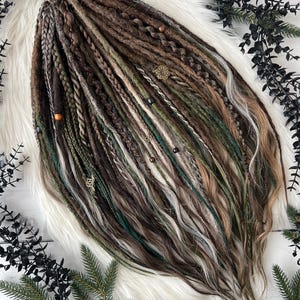 May include: Handmade dreadlocks in shades of brown, green, and blonde, with decorative beads and charms. The dreads are arranged on a white faux fur surface, with dark green foliage accents.