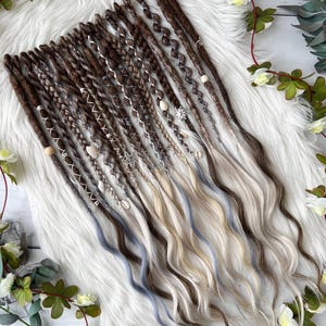 May include: A collection of brown, blonde, and blue synthetic dreadlocks. Some dreadlocks are adorned with beads, shells, and silver charms. The dreadlocks are arranged on a white, fluffy surface and decorated with greenery and white flowers.