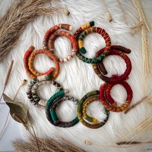 Op de afbeelding: Een set van tien kleurrijke, handgeweven armbanden van stof. De armbanden zijn gemaakt in verschillende kleuren, waaronder rood, oranje, geel, groen en bruin. De armbanden zijn in een cirkel op een witte achtergrond gerangschikt.