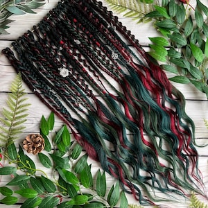 Puede incluir: Un conjunto de rastas en negro, rojo oscuro y verde azulado. Algunas rastas están adornadas con abalorios plateados. Las rastas están dispuestas sobre una superficie clara con vetas de madera, rodeadas de follaje verde y piñas.