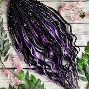 May include: A bundle of black, purple, and lavender synthetic dreadlocks with wavy ends. The dreadlocks are decorated with silver-colored charms. The hair is arranged on a light wood-grain surface, with pink flowers and green leaves in the background.
