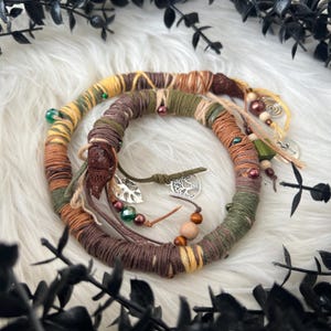 Puede incluir: Una envoltura de pelo trenzada marrón, verde y amarilla con cuentas de madera, encantos y un colgante de árbol plateado.