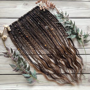May include: A set of brown and blonde synthetic dreadlocks with beads and charms. The dreadlocks are hanging down and are arranged in a fan shape.