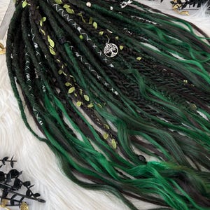 May include: A set of dark green and black dreadlocks with bright green tips. The dreads are adorned with small green leaves, silver accents, and a silver tree of life charm. The dreadlocks are arranged on a white, fluffy surface.