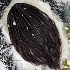 May include: Dark brown dreadlocks with lighter brown highlights and silver accents. The dreadlocks are styled with decorative silver charms and beads. The hair is arranged on a white faux fur surface, with dark green and white floral accents.
