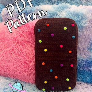 May include: A dark brown, rectangular crochet item with colorful pom-poms scattered across its surface. The text "PDF Pattern" is at the top, and the logo "Cosmic Crafts and Crochet" is at the bottom. The item is set against a backdrop of fluffy pink and blue pillows.