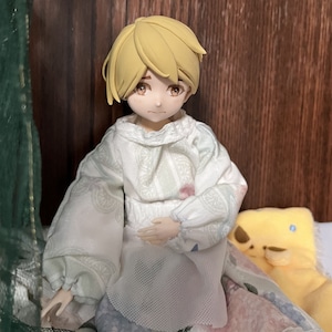 May include: A doll with short, blonde hair wearing a white, floral-patterned dress with a lace collar. The doll is sitting on a bed with a yellow pillow.
