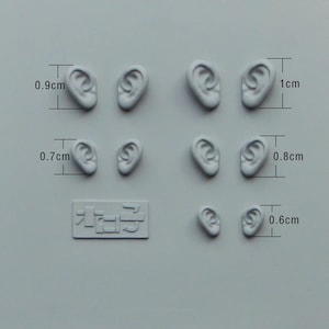 May include: A collection of small, light gray ear models in various sizes, ranging from 0.6 cm to 1 cm in height. The models are arranged on a light gray surface, with a small rectangular plaque with Japanese text.