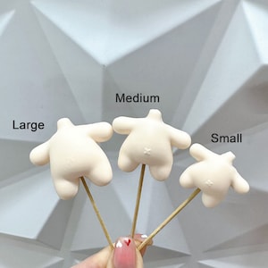 May include: Three off-white, torso-shaped cake toppers on wooden skewers, labelled "Large", "Medium", and "Small". The toppers have arms and a small flower detail on the back. The background is a white, geometric pattern.