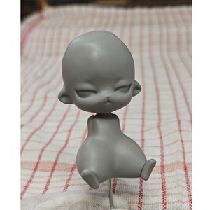 Puede incluir: Una cabeza y un cuerpo de muñeca grises sin pintar con una nariz pequeña y los ojos cerrados. La muñeca está sentada sobre una superficie a cuadros blancos y rojos.